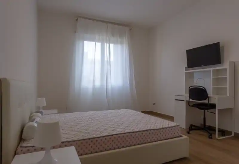 Dormitorio luminoso y de mobiliario sencillo con cama doble, lámparas de mesilla y un escritorio con TV en la pared. La luz natural filtrada por las cortinas aporta una atmósfera tranquila y neutra, ideal para alquileres de corta estancia.
