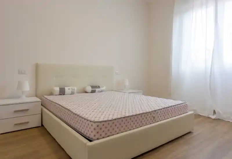 Dormitorio luminoso y minimalista con una gran cama tapizada, mesitas a juego y luz natural suave desde una ventana con cortina — limpio, moderno y listo para preparar.