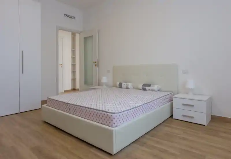Dormitorio luminoso y minimalista con una amplia cama tapizada, dos mesitas de noche y suelo de madera claro; tonos neutros y decoración sencilla que transmiten calma para anuncios.