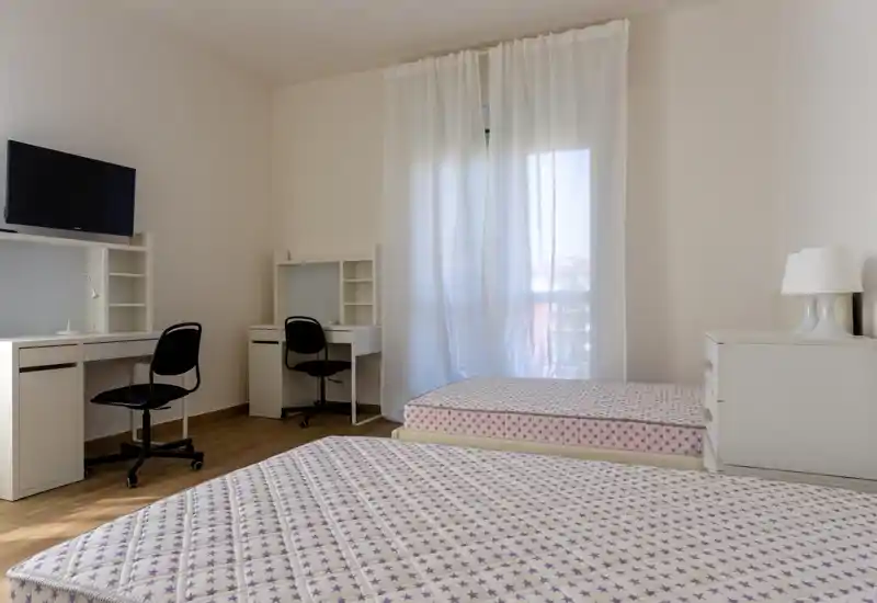 Dormitorio luminoso y minimalista con dos camas individuales, muebles blancos sencillos y una gran ventana con cortinas translúcidas que dejan pasar la luz natural.