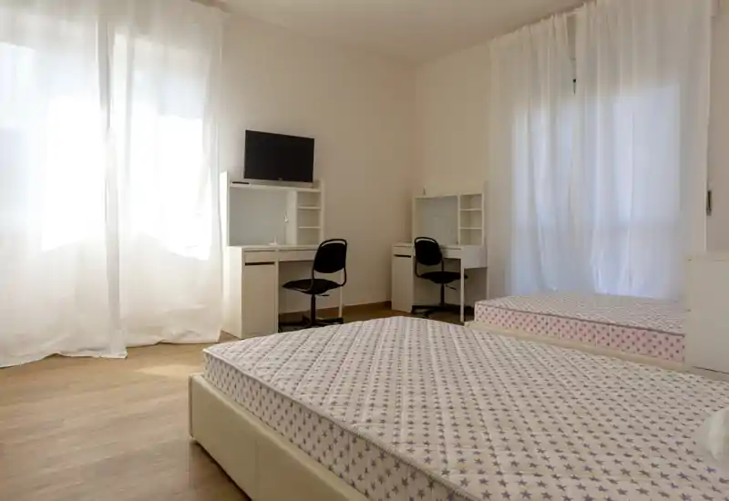 Habitación luminosa con dos camas individuales, escritorios y sillas a juego, y grandes ventanas con cortinas translúcidas que aportan mucha luz natural.