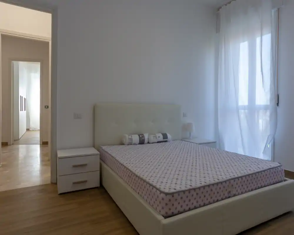 Camera da letto luminosa e minimal con letto matrimoniale, due comodini e luce naturale da una finestra con tenda: linee pulite e toni neutri creano un’atmosfera tranquilla.