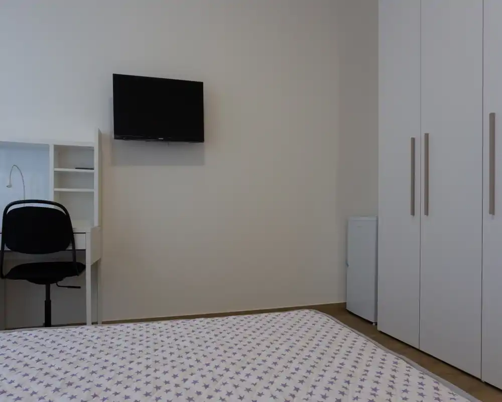 Angolo di camera da letto minimalista con copriletto a fantasia in primo piano, TV a parete, scrivania con sedia e un alto armadio bianco — luminoso, pulito e funzionale.