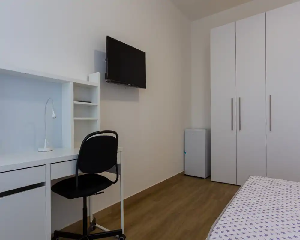 Angolo di camera da letto moderno e minimal con letto singolo visibile, armadio bianco, scrivania e TV a parete — luminoso, pulito e funzionale per soggiorni brevi.