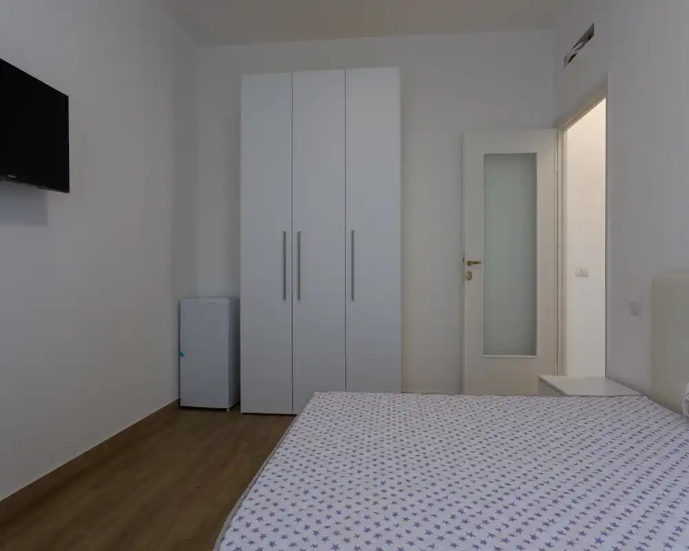 Camera da letto moderna e minimal con ampio letto in primo piano, armadio bianco e arredi essenziali. Pavimento effetto legno e tonalità neutre per un'atmosfera tranquilla.