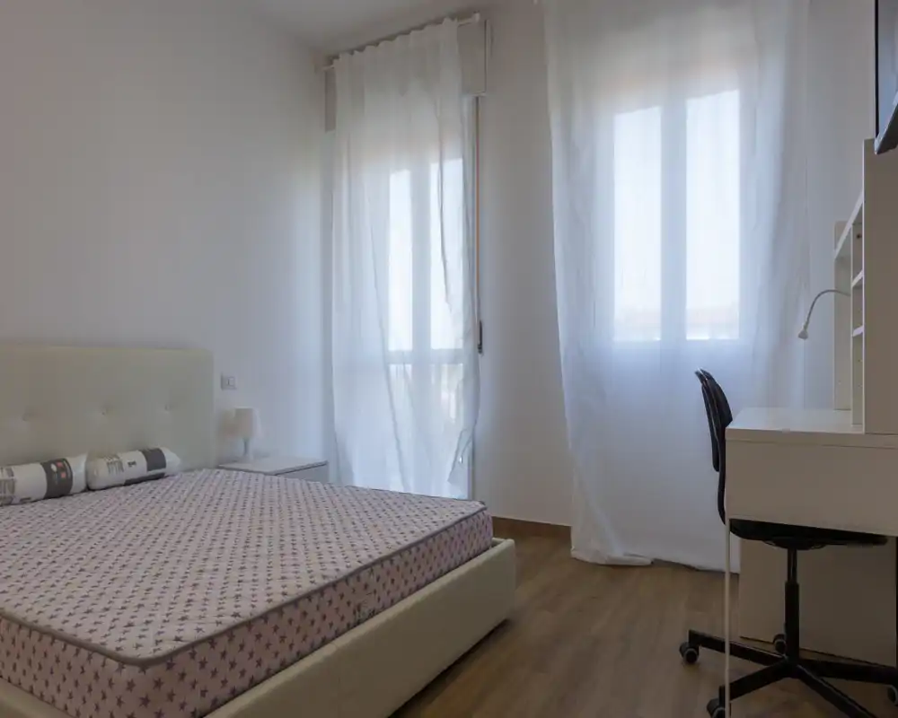 Camera da letto luminosa e minimale con letto matrimoniale, tende leggere e una scrivania vicino alla finestra. Arredo neutro e pavimento effetto legno per un’atmosfera moderna e rilassante.