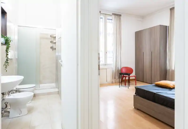 Camera da letto moderna e luminosa con letto visibile, armadio e finestra che illumina la stanza; arredo semplice e accogliente con copriletto scuro e cuscino accentato.