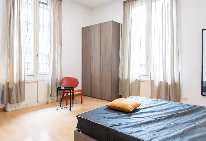 Dormitorio luminoso y minimalista con cama grande, armario de madera y ventanas altas que dejan entrar abundante luz natural.