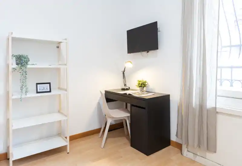 Angolo studio luminoso e minimal con scrivania, sedia, lampada e TV a parete vicino a una finestra con tenda leggera — ideale per un monolocale o smart working.