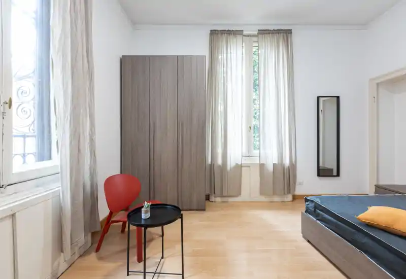 Camera da letto luminosa e minimal con letto basso visibile, armadio e finestra alta che diffonde luce naturale. Linee pulite e tonalità neutre creano un’atmosfera tranquilla e contemporanea, ideale per affitti brevi.