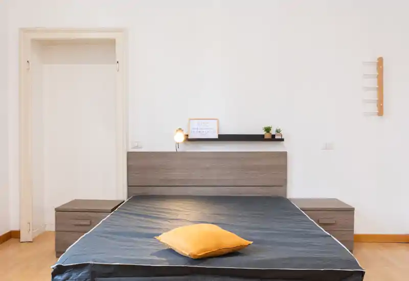 Dormitorio luminoso y minimalista con cama doble centrada, mesitas a juego y pequeños detalles decorativos (lámpara, plantas). La habitación está limpia, bien iluminada y fotografiada de frente, apta para anuncios de alquiler/venta.