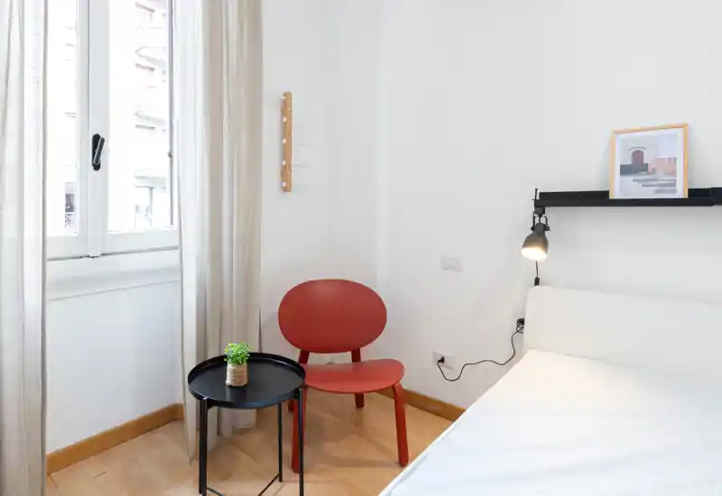 Rincón de dormitorio luminoso y minimalista con cama blanca, lámpara de pared y una pequeña zona de asiento con silla roja y mesita negra redonda junto a la ventana.