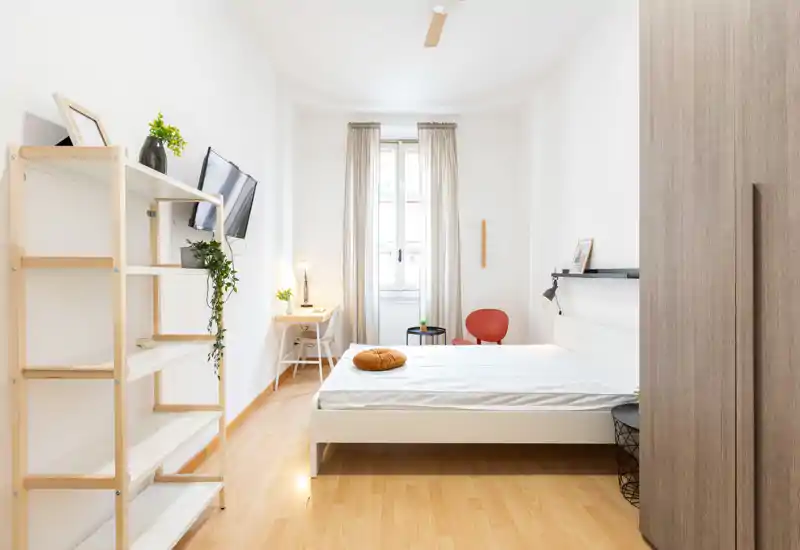 Dormitorio luminoso y minimalista con cama blanca, suelo de madera clara, un escritorio junto a la ventana y detalles cuidados como un cojín redondo y plantas.
