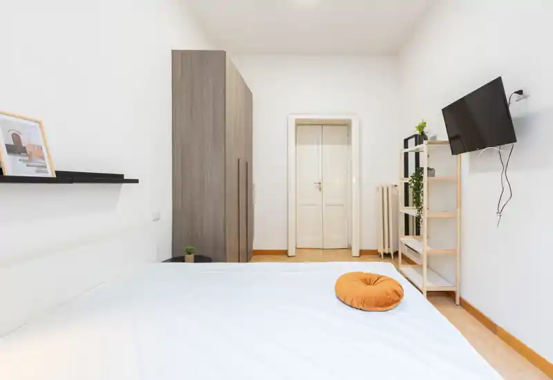 Dormitorio luminoso y minimalista con una gran cama en primer plano, armario y estanterías, y TV de pared; perfecto para anuncios que destaquen un espacio para dormir limpio y aireado.