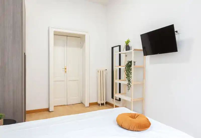 Dormitorio luminoso y minimalista con la cama en primer plano, paredes neutras, suelo de madera, TV en la pared y estantería con plantas — habitación limpia y acogedora para anuncios.