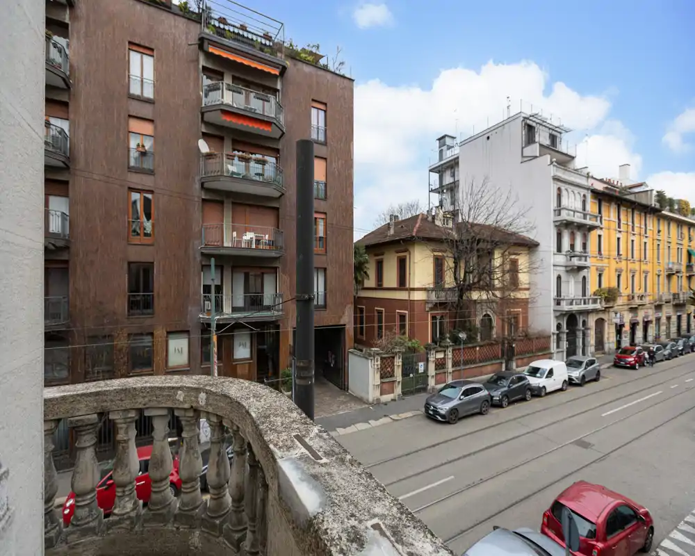 Balcone con vista su una strada cittadina, auto parcheggiate e edifici residenziali vicini; luce diurna e cielo sereno.