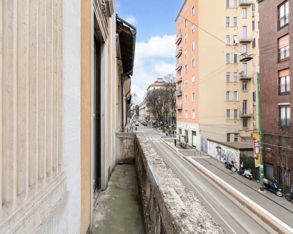 Balcone stretto e soleggiato con vista sulla strada cittadina, edifici residenziali e traffico leggero — perfetto per un caffè mattutino.