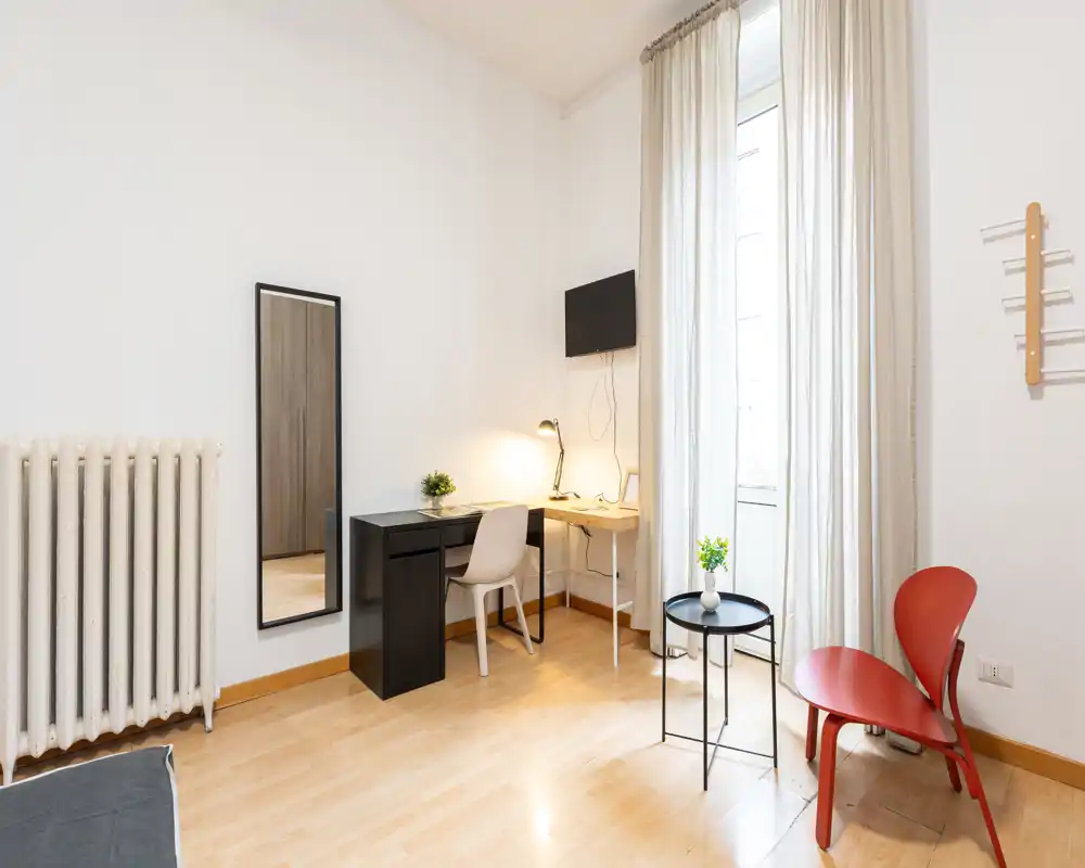 Angolo di camera luminoso e arredato in modo essenziale con scrivania, TV a parete e area salotto vicino a una grande finestra con tende leggere.