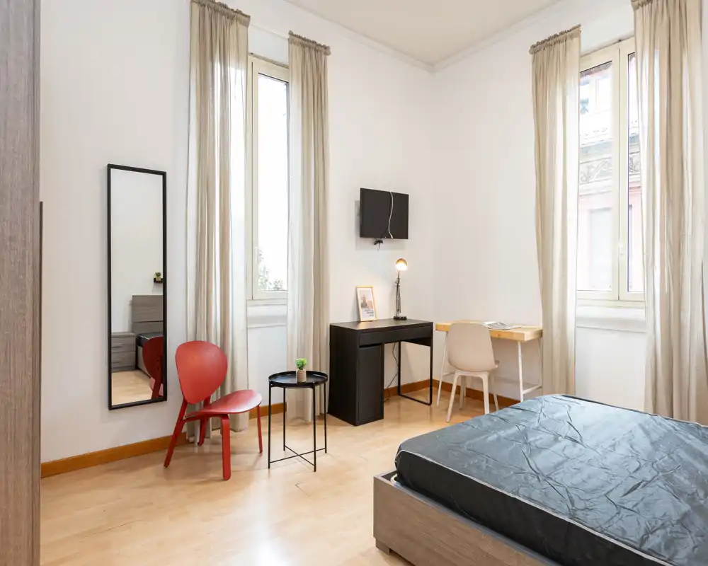 Camera da letto luminosa e arredata in modo semplice con letto visibile, due finestre alte con tende leggere, una scrivania e un piccolo angolo seduta — ideale per una persona o soggiorni brevi.