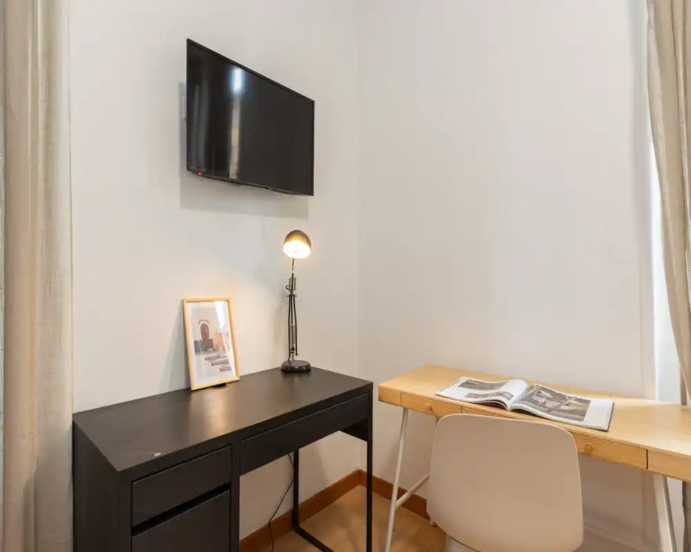 Angolo living/lavoro ordinato con TV a parete, una scrivania scura, un tavolo in legno con sedia e una lampada da tavolo — foto nitida e luminosa che mostra uno spazio compatto e funzionale.