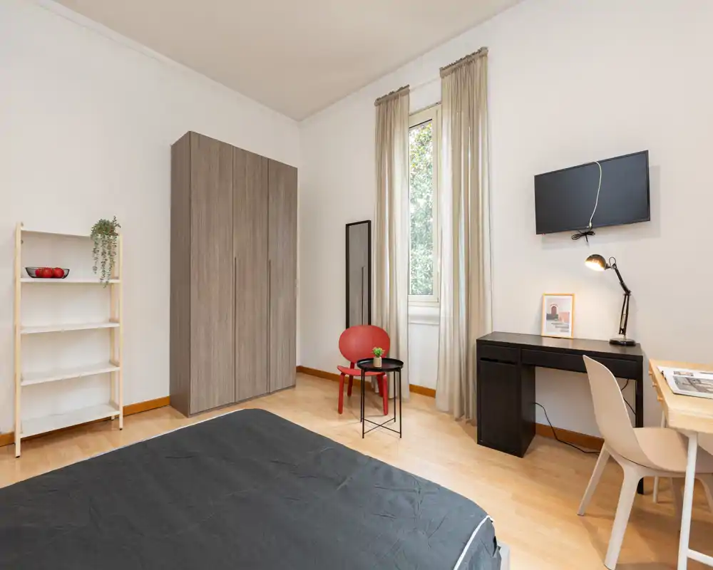 Camera da letto luminosa e ben arredata con letto visibile, armadio, scrivania e area relax vicino a una finestra alta che fa entrare luce naturale. Pavimento in legno chiaro e tonalità neutre donano un'atmosfera moderna e tranquilla.
