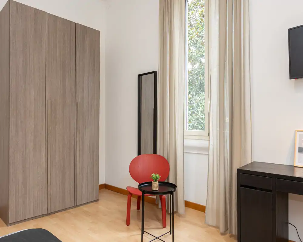 Angolo di camera da letto luminoso e minimal: grande armadio, finestra con tende leggere, una piccola sedia rossa e un tavolino; stile moderno e pulito.
