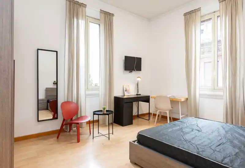 Camera da letto luminosa e arredata in modo semplice con letto visibile, due finestre alte con tende leggere, una scrivania e un piccolo angolo seduta — ideale per una persona o soggiorni brevi.