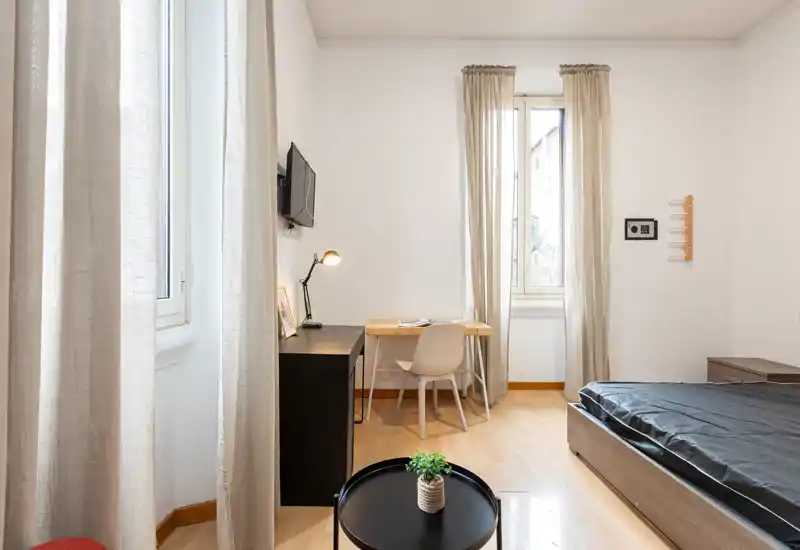 Dormitorio luminoso y minimalista con cama visible, escritorio y luz natural de ventanas altas; tonos neutros y decoración sencilla que crean un ambiente moderno y relajado.