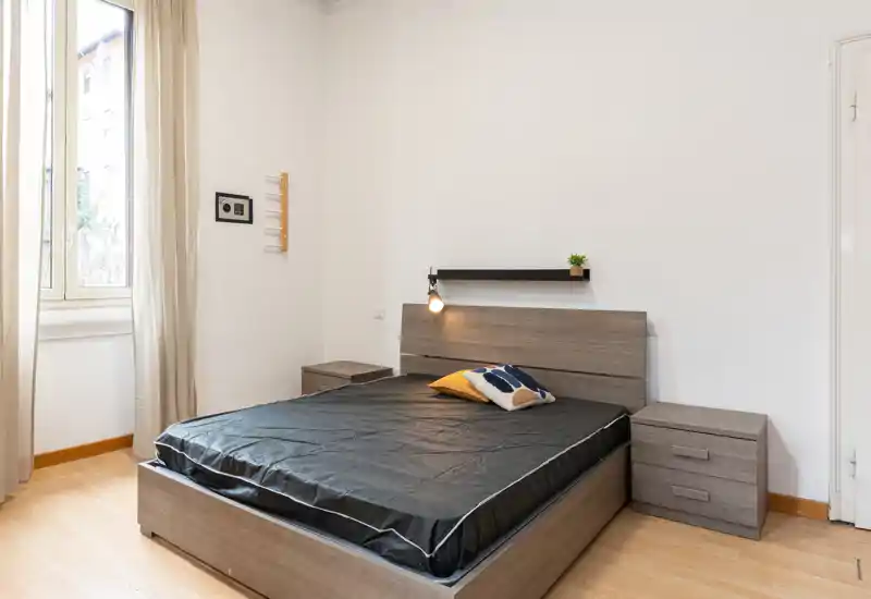 Dormitorio moderno y luminoso con cama doble, dos mesitas de noche y una gran ventana que aporta luz natural. Decoración sencilla y neutra con lámpara de cama y cojín decorativo.