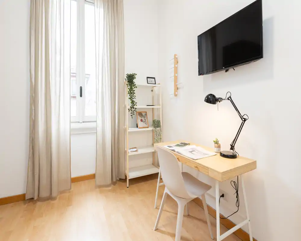 Pequeño rincón de dormitorio luminoso habilitado como zona de estudio: escritorio de madera con lámpara y silla bajo televisor en la pared, suelo de madera claro, estantería y cortinas translúcidas que dejan entrar la luz natural.