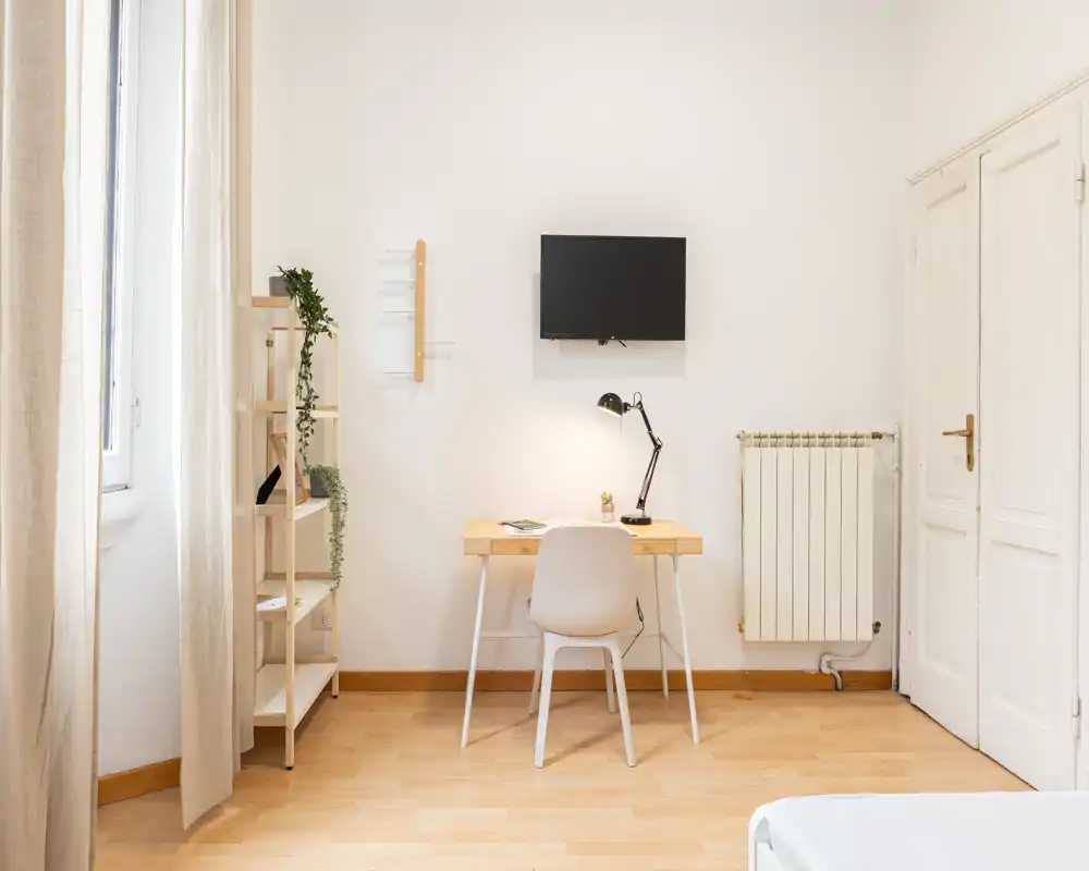 Rincón de dormitorio luminoso y minimalista con un pequeño escritorio, silla y televisor de pared. La luz natural y los detalles de madera crean un área de trabajo tranquila junto a la cama.
