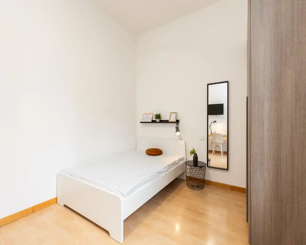 Dormitorio individual luminoso y minimalista con cama blanca, estante junto a la cama, espejo y armario. Suelo de madera y decoración sencilla que crean un espacio acogedor y funcional.