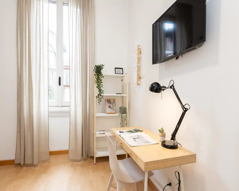 Rincón de salón luminoso y minimalista con escritorio de madera, silla y televisor en la pared; cortinas ligeras y pequeñas plantas crean un acogedor espacio de trabajo.