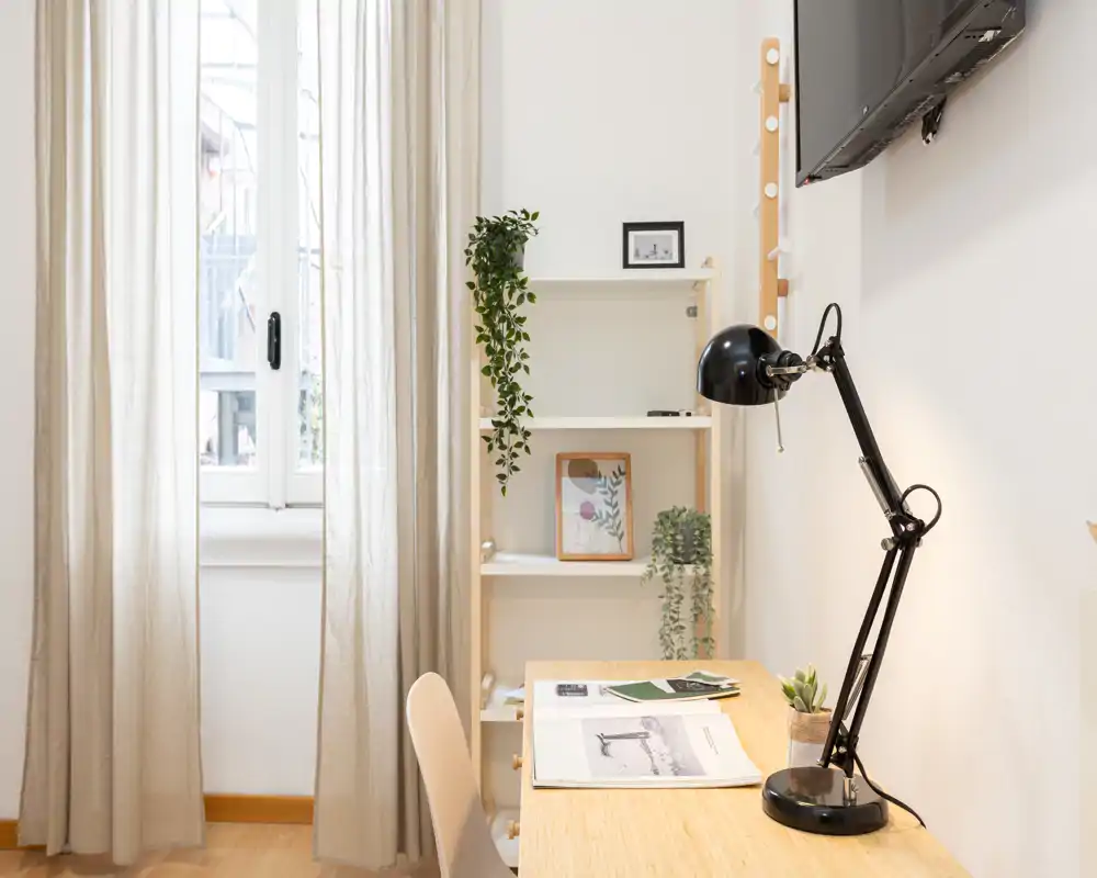 Espacio de trabajo luminoso y minimalista junto a la ventana con escritorio de madera, lámpara de escritorio negra ajustable y plantas decorativas — ideal para trabajar desde casa.