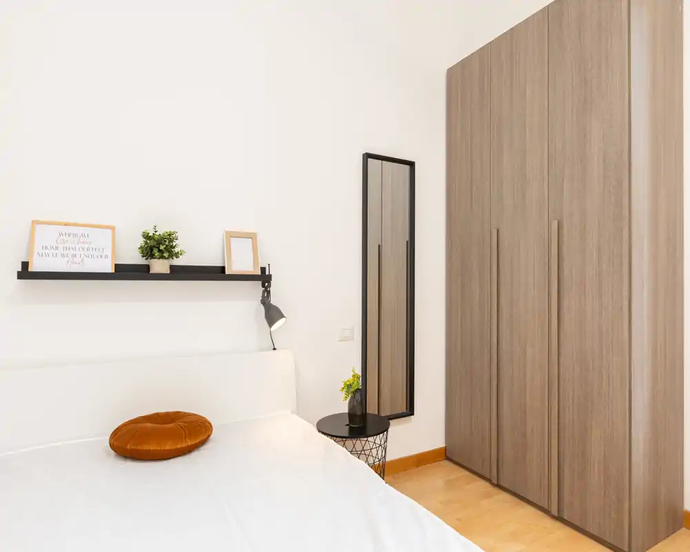 Dormitorio luminoso y minimalista con cama doble, lámpara de mesita, pequeña mesa auxiliar y un armario alto de madera — líneas limpias y luz natural suave.