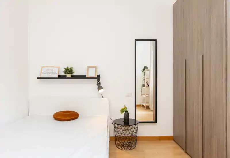 Dormitorio minimalista y luminoso con estructura de cama blanca, cojín redondo color ocre, lámpara de mesita y un espejo alto junto a un gran armario — limpio, aireado y bien iluminado.