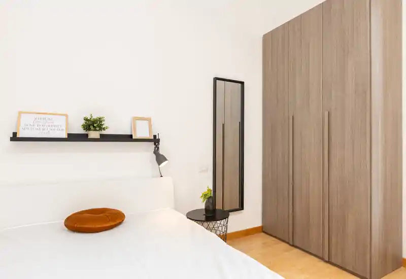 Dormitorio luminoso y minimalista con cama doble, lámpara de mesita, pequeña mesa auxiliar y un armario alto de madera — líneas limpias y luz natural suave.