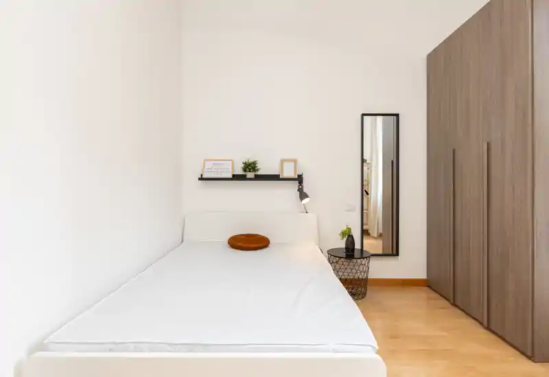 Dormitorio minimalista con una cama individual bien hecha, paredes blancas, suelo de madera y un gran armario empotrado. Detalles sencillos como un cojín redondo, una pequeña estantería y un espejo de cuerpo entero crean un ambiente limpio y luminoso.