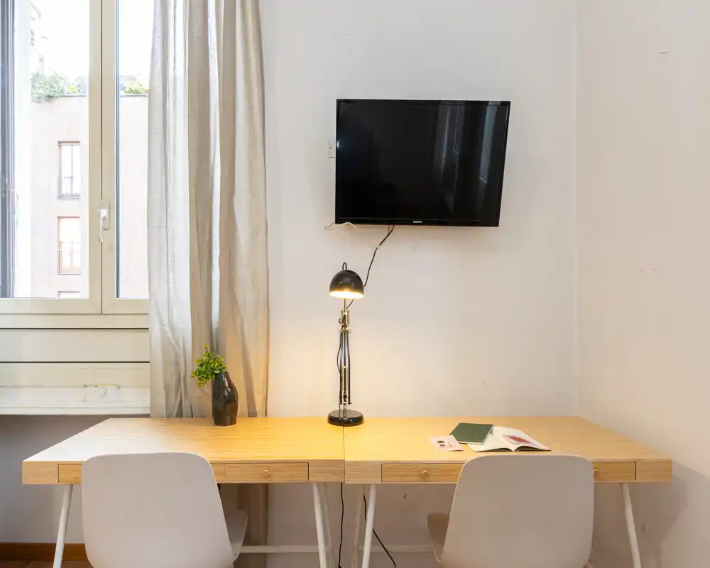 Área de trabajo luminosa y minimalista con una mesa doble de madera, dos sillas, una lámpara de escritorio y una TV mural junto a una gran ventana.