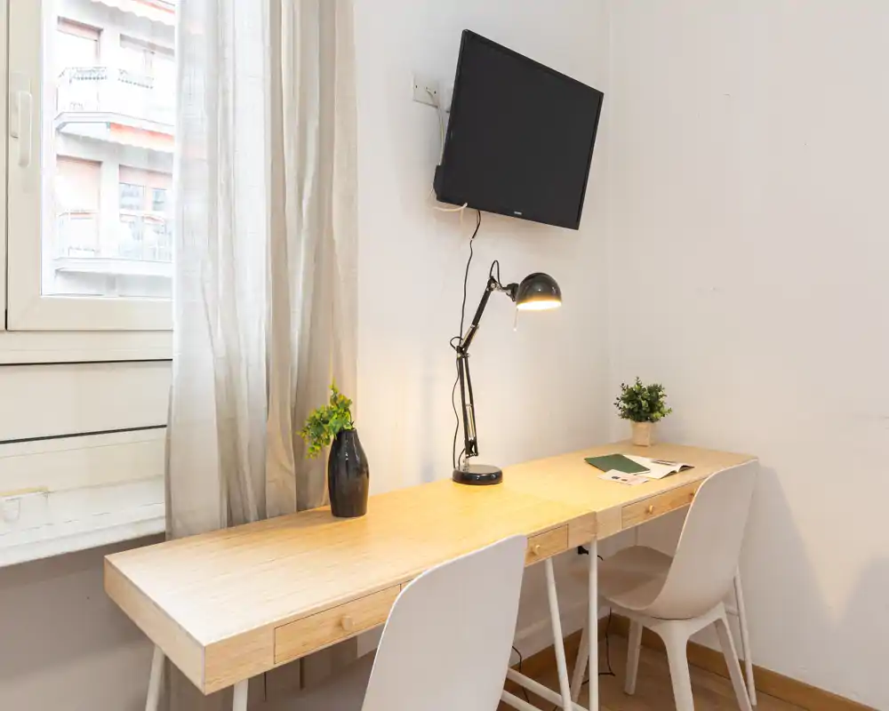 Espacio de trabajo luminoso con una larga mesa de madera, dos sillas, una lámpara de tarea y plantas decorativas junto a la ventana — ideal para estudiar o trabajar en remoto.