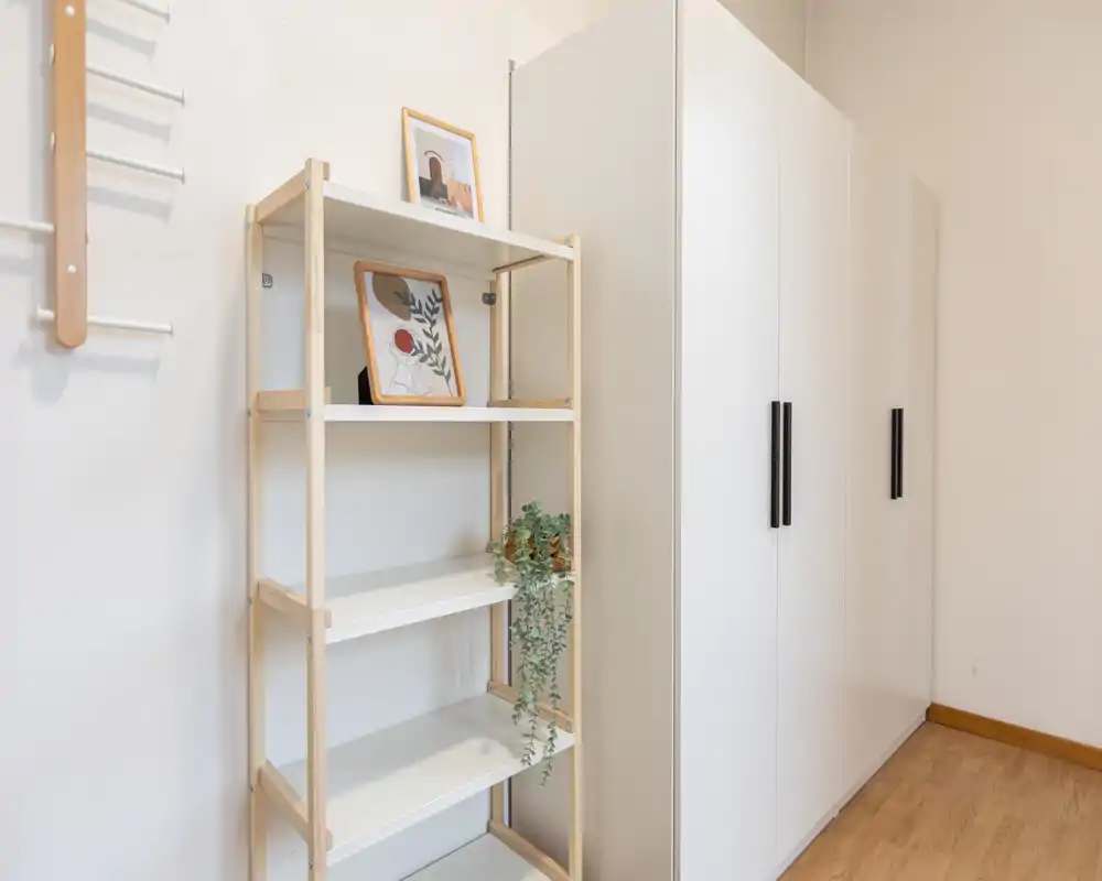 Rincón de dormitorio minimalista con un armario alto blanco y una estantería de madera clara decorada con cuadros y una planta colgante. Limpio, luminoso y de estilo sencillo — perfecto para mostrar opciones de almacenamiento y decoración.