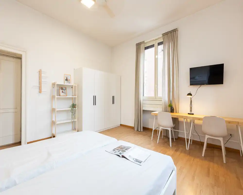 Dormitorio luminoso y minimalista con dos camas individuales juntas, un armario y un escritorio largo junto a la ventana, ideal para trabajar desde casa.