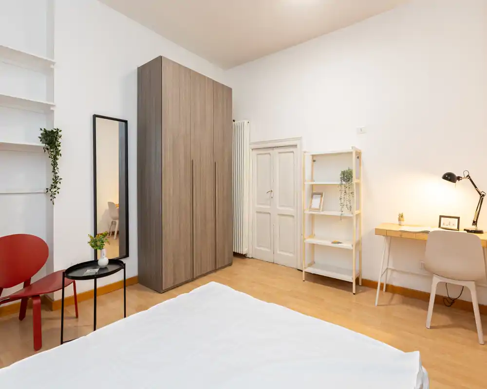 Dormitorio luminoso y minimalista con la cama visible en primer plano, un armario alto de madera, escritorio con lámpara y estantería ordenada — espacioso, bien iluminado y de estilo sencillo.