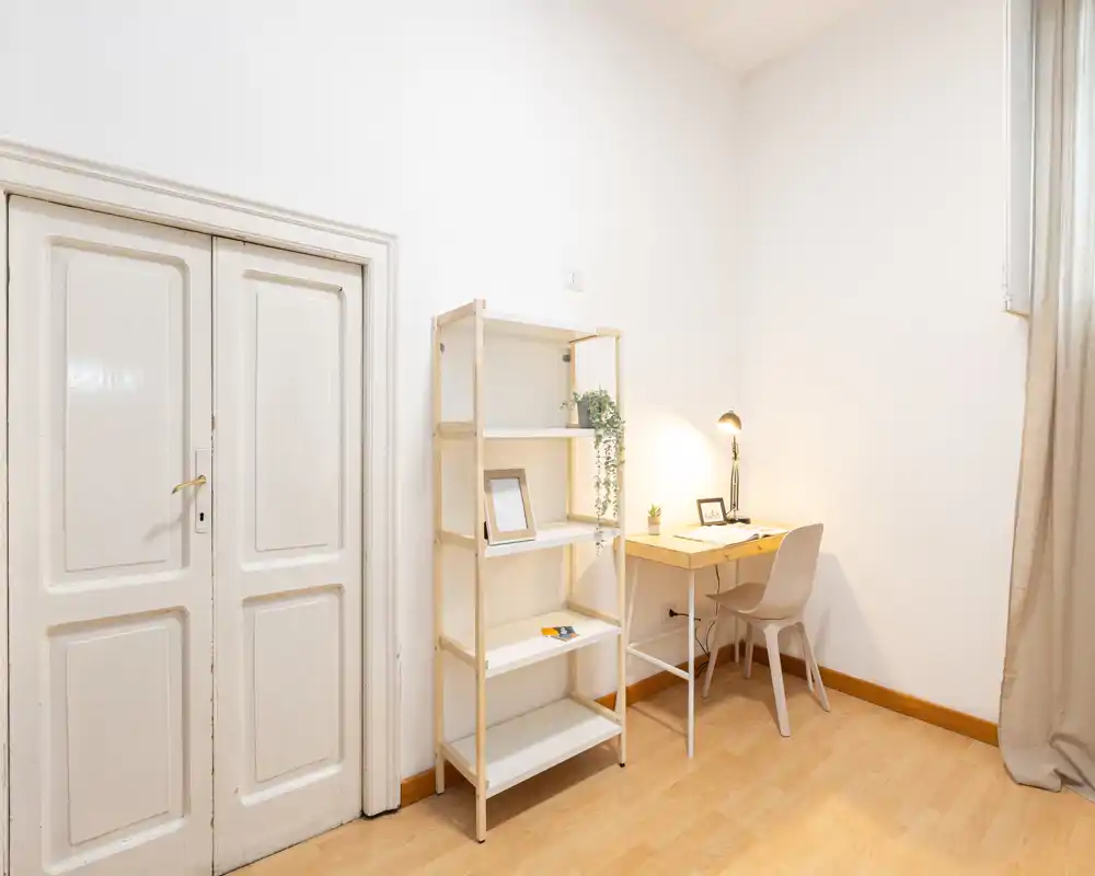 Rincón de estudio luminoso y minimalista con escritorio de madera, silla, lámpara y estantería—ideal para teletrabajo o lectura.
