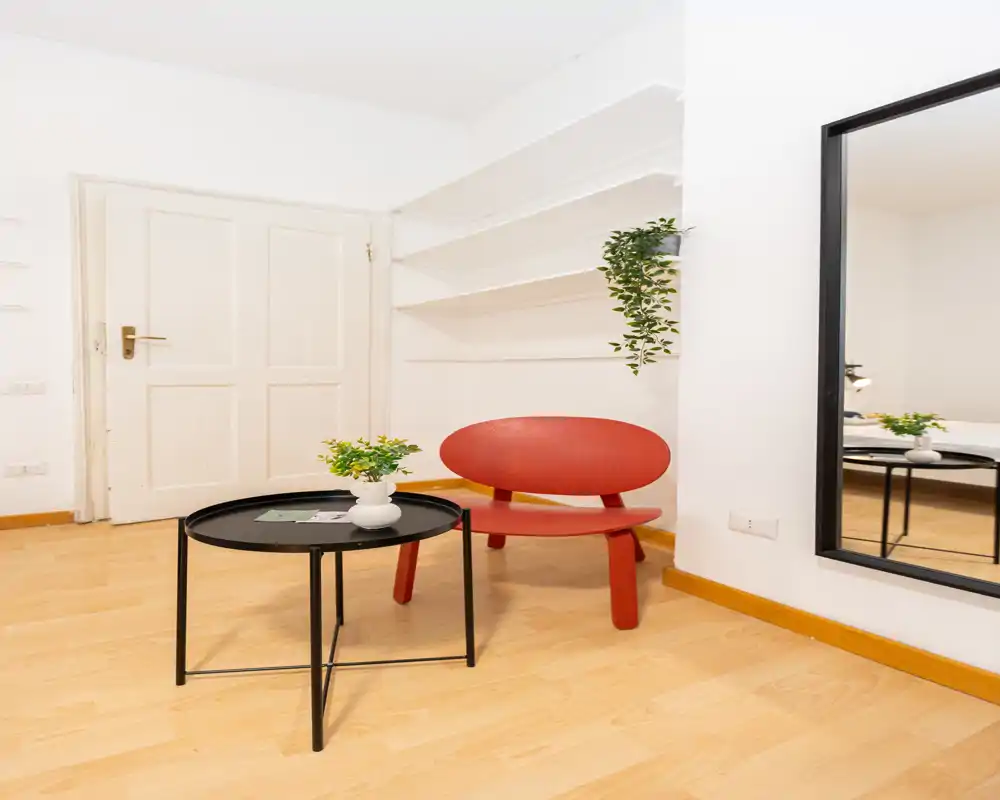 Pequeña zona de estar moderna con sillón rojo de diseño, mesa de café redonda negra y plantas decorativas sobre pared blanca luminosa: estilo limpio y minimalista perfecto para un piso compacto.