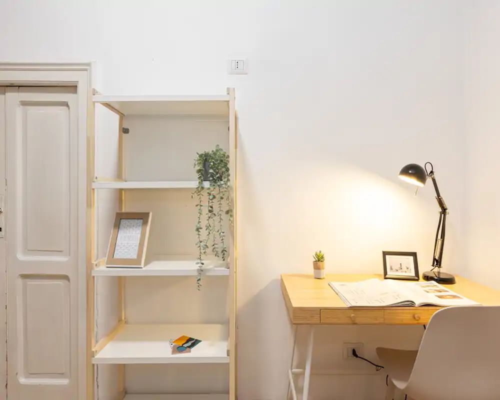Área de estar luminosa y minimalista con escritorio de madera y estantería — ideal como pequeña oficina en casa o rincón de estudio, con lámpara de escritorio, planta y marco decorativo.