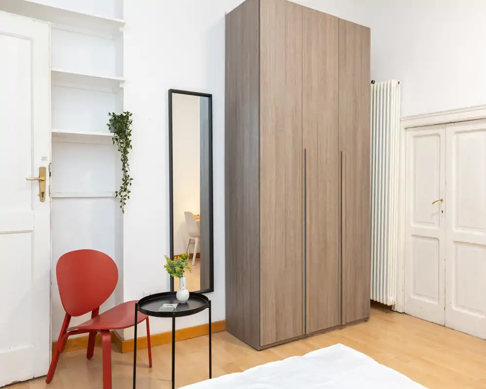 Rincón de dormitorio luminoso y de estilo sencillo con un gran armario de madera, una mesa auxiliar redonda, una silla roja como acento y un espejo estrecho en la pared; se ve parcialmente la ropa de cama en primer plano.