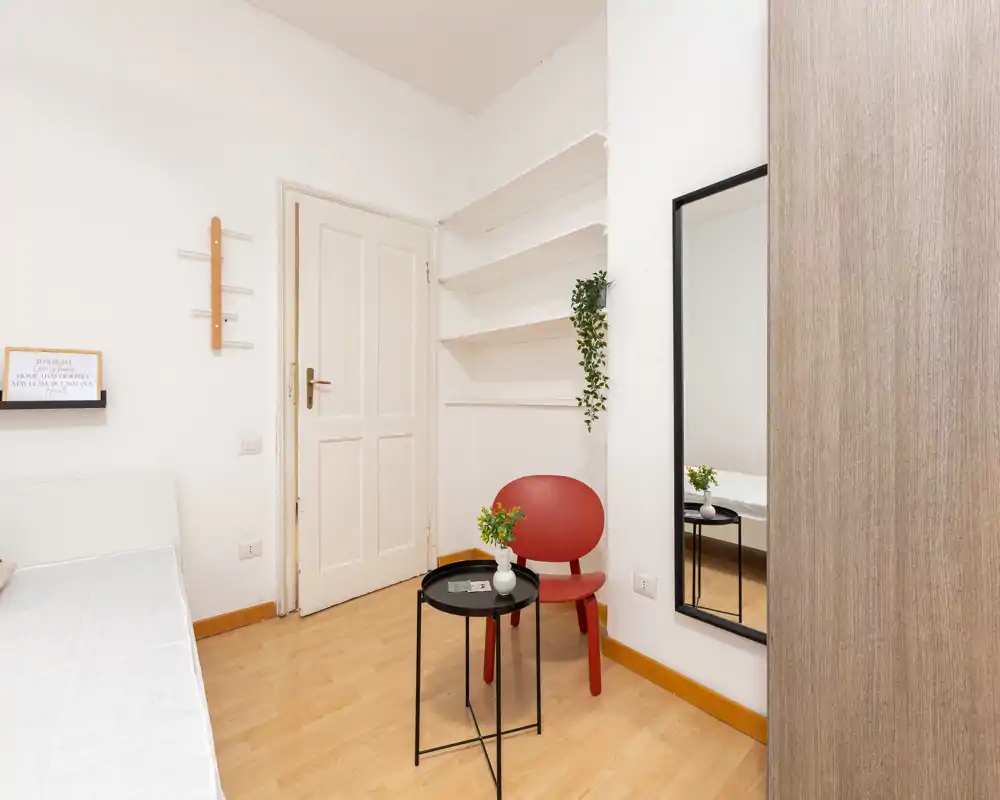 Rincón de dormitorio luminoso y minimalista con el borde del colchón visible, una silla roja, una mesa redonda pequeña y un espejo que aporta profundidad. Estilo limpio y moderno ideal para anuncios de alquiler.