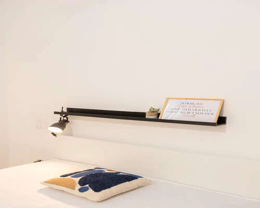 Imagen luminosa y minimalista de dormitorio con cabecero, estante de pared con decoración y cama arreglada con cojín decorativo, perfecta para anuncios modernos.