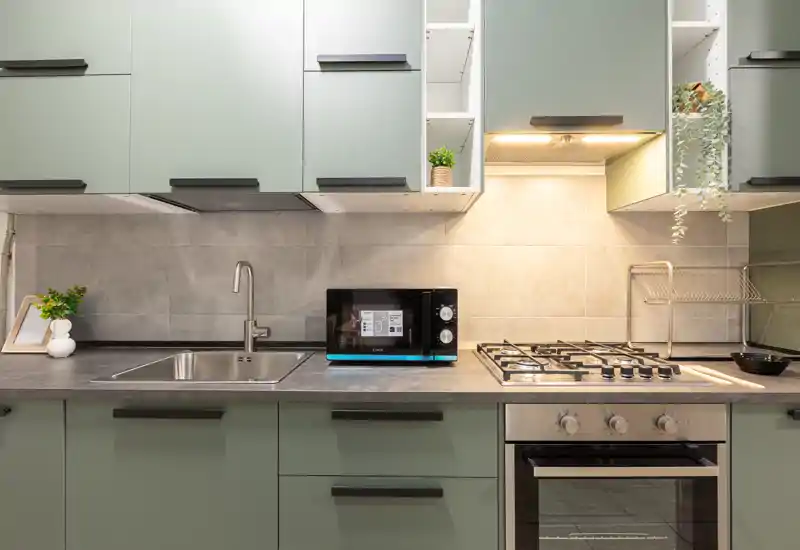 Cucina moderna e compatta con pensili verde salvia, lavello in acciaio, piano cottura a gas e forno incassato. Piano di lavoro ben illuminato con microonde e piante che aggiungono un tocco di casa.